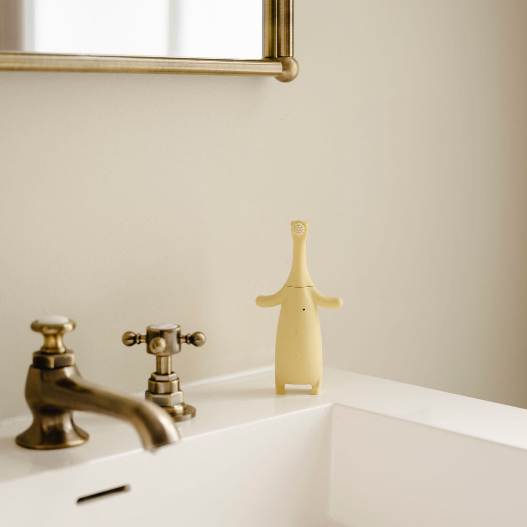 
Brushi Bear™ in the Bathroom next to a sink with gold faucet and against a beige wall.
