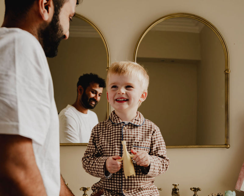 Child in checkered pajamas sitting on a bathroom counter with a man, reflected in a mirror, using Brushi Bear toothbrush